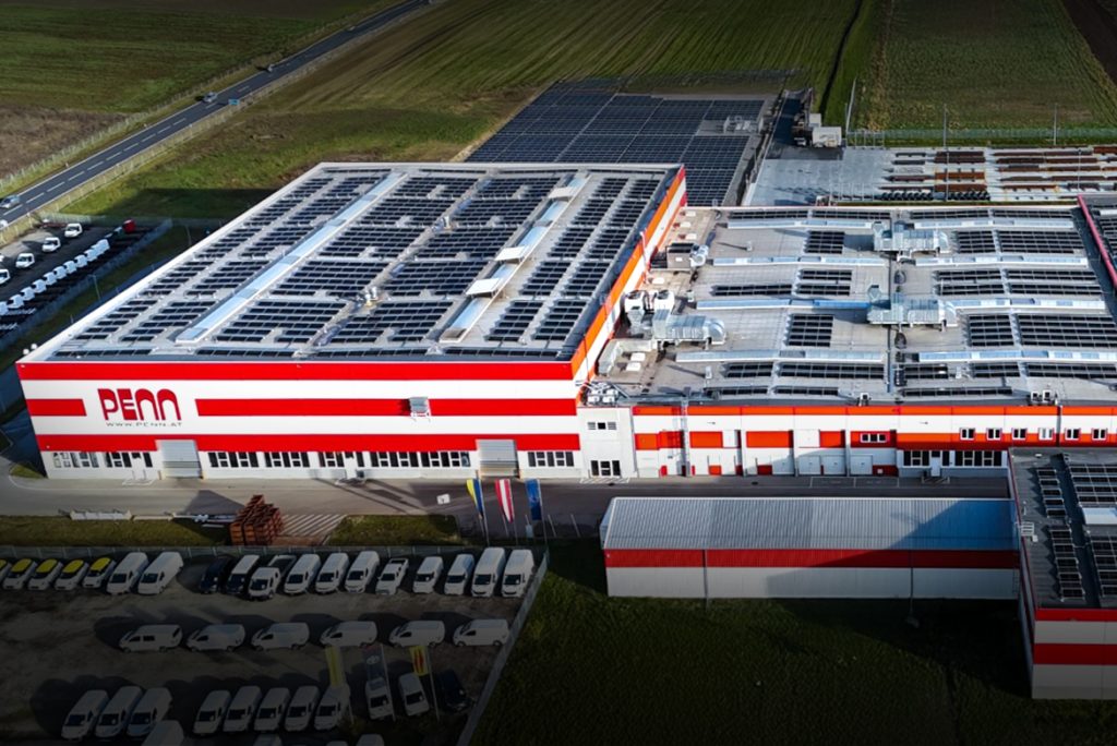 Luftaufnahme des PENN GmbH Industriekomplexes in Stratzdorf mit mehreren modernen Produktionsgebäuden und rot-weißen Fassadenelementen, umgeben von Parkplätzen und landwirtschaftlichen Flächen.